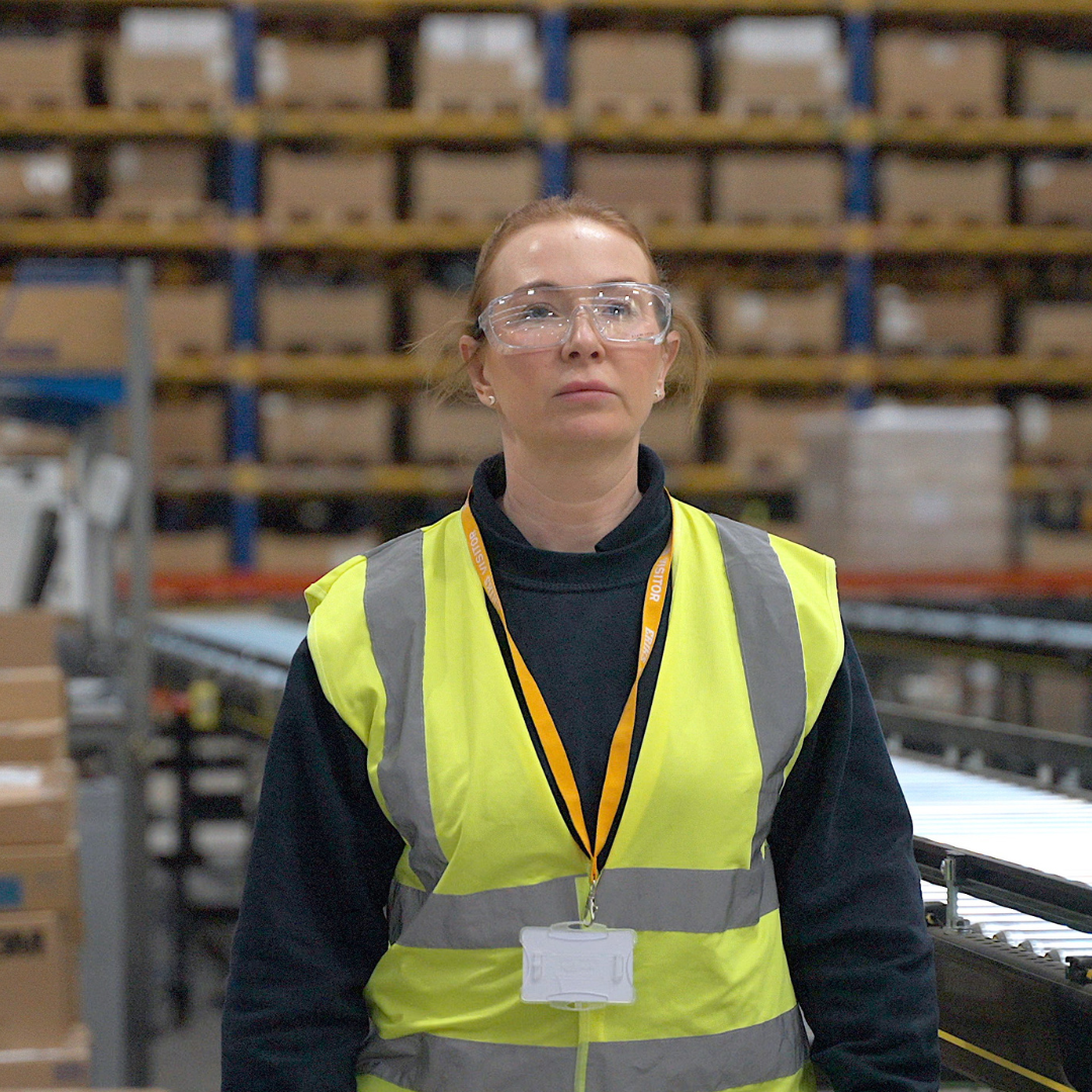 A regal standing woman in a high-vis and PPE in a big warehouse looking up